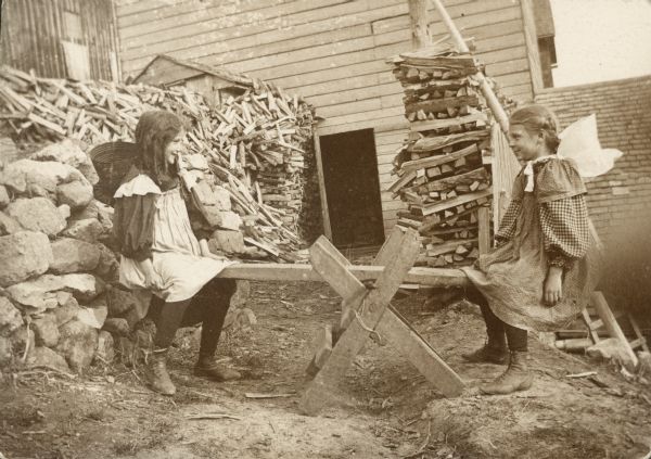 Two young girls play on a seesaw (teeter totter) in a yard near stacks of fuel wood.