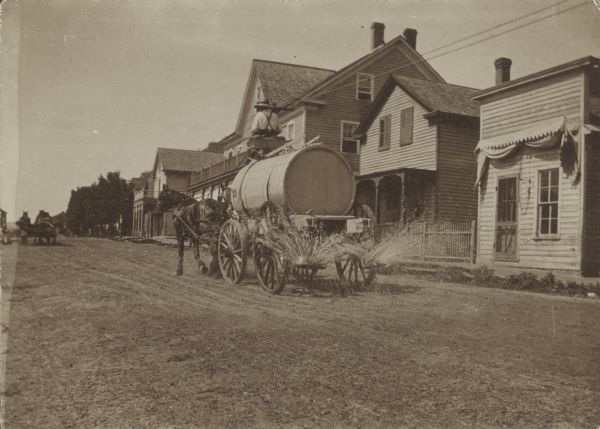 A man with two horses pulling a street waterer down a dirt road. When Main Street was a dirt road, the local merchants paid to have it sprinkled during the summer to keep the dust down.
