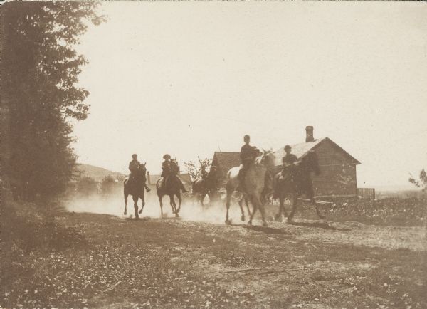 Five men ride horses and kick up a cloud of dust along a country road. A small homestead is in the background.