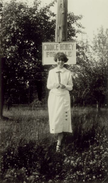 Outdoor portrait of a young dark-haired women standing in a field of flowers before a sign attached to a telephone pole. The sign reads: "Choice Honey For Sale."