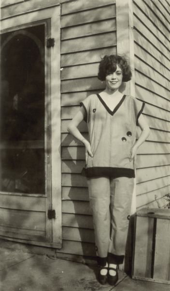 Outdoor portrait of a young dark-haired woman standing in front of the of a building near a screen door.