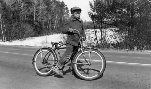 A young Menominee Indian (Charles ?) pauses briefly for a photograph while straddling his bicycle. He is stopped on the road with the snow-covered Wolf River behind him.