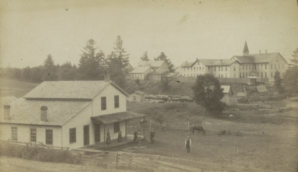 Farmer's house with rear of the government Contract School on the Menominee reservation in the distance. The Keshena Creek flows between the farmhouse and the school. A group of people stand on and near the porch of the house.