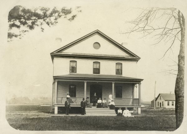 Exterior of Keshena Boardinghouse with a group of men, women and children positioned on the front porch.