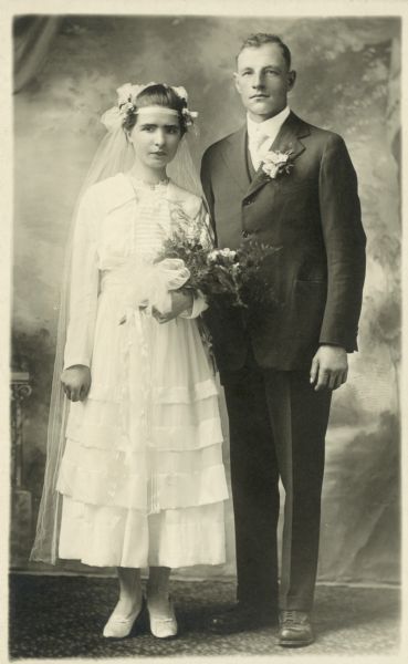 Photographic postcard of a studio portrait in front of a painted backdrop of Mr. & Mrs. Louis Buechner on their wedding day. She wears a traditional wedding gown and holds a bouquet of flowers, and he has a boutonnière pinned to his lapel.