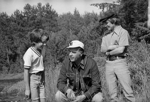 Nelson takes time out during a campaign trip to talk with two boys. Dressed casually, Nelson is shown squatting down in a marsh area between the boys who are standing on either side of him.