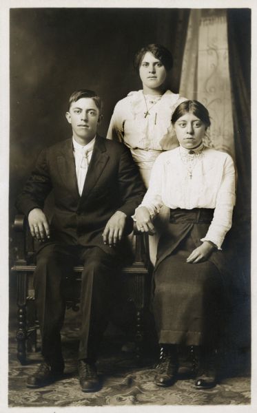 Photographic postcard of a studio group portrait of (left to right) George Staty, Mary Staty Marx and Margaret Staty. They are standing in front of a painted backdrop.