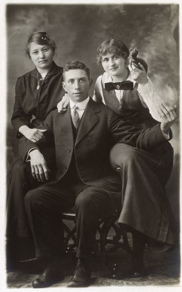 Photographic postcard of a studio group portrait of (left to right) " Elizabeth "Lizzie" Schoepp, Anton Joseph Dahmen, and Eva Dahmen Bowar. Eva holds a toy stuffed animal in her left hand. They are standing in front of a painted backdrop. Eva is Anton's younger sister.