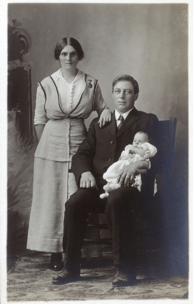 Photographic postcard of a studio portrait of Elizabeth Dohn Staty, her husband Wolfgang Staty and their infant Martha Staty. They are standing in front of a painted backdrop.