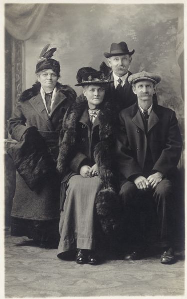 Photographic postcard of a studio group portrait of two men and two women, standing in front of a painted backdrop. They are all wearing hats.