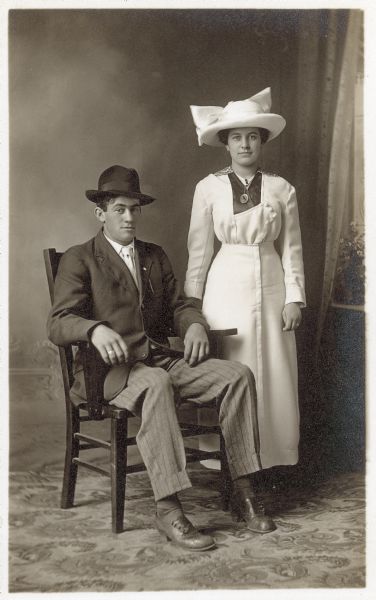 Photographic postcard of a studio portrait of (brother and sister?) Mike Endres and Katie Endres Mick, in front of a painted backdrop. They are both wearing hats.
