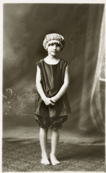 Photographic postcard of a studio portrait of an unidentified young girl wearing a bathing suit and cap, standing in front of a painted backdrop.