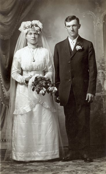 Photographic postcard of a studio portrait of a couple on their wedding day. She wears a traditional wedding gown and holds a bouquet of flowers and he has a boutonniere pinned to his lapel. They are standing in front of a painted backdrop.
