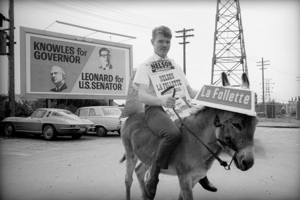 An unidentified man wearing a sandwich board supporting Bronson La Follette for Governor and Gaylord Nelson for United States Senator is seated on a donkey with a La Follette sign over its ears during the "Whistle Stop" campaign. In the background is a billboard advertising Knowles for Governor and Leonard for U.S. Senator.