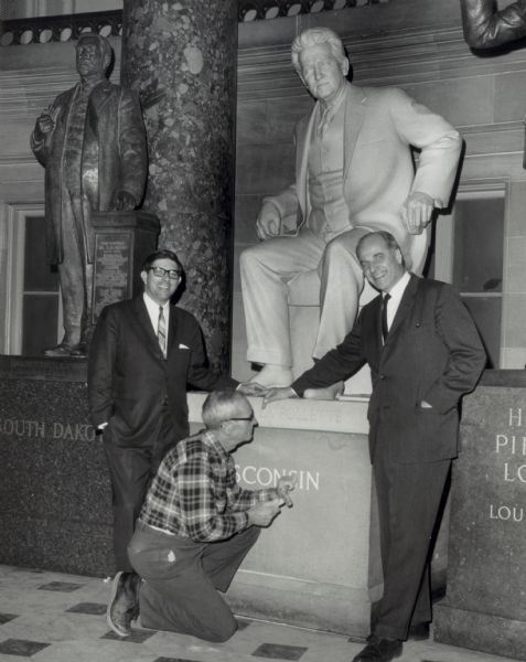 Nelson and Bronson La Follette stand next to the Robert M. La Follette statue in the United States Capitol, while a man poses at the base to carve the word Wisconsin.