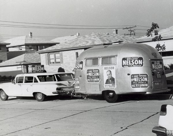A car and a trailer covered with "Gaylord Nelson for U.S. Senator" posters sits parked on a neighborhood street.
