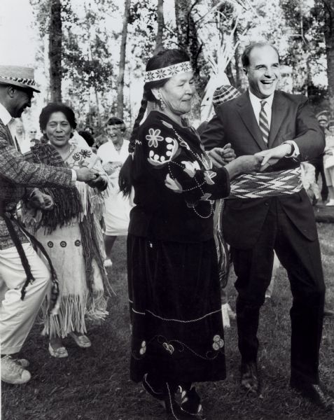 Nelson dances outside with an older Ojibwa woman wearing a traditional beaded dress and headband. Nelson is dressed in a suit and wears a beaded sash; others are also pictured dancing in the background.