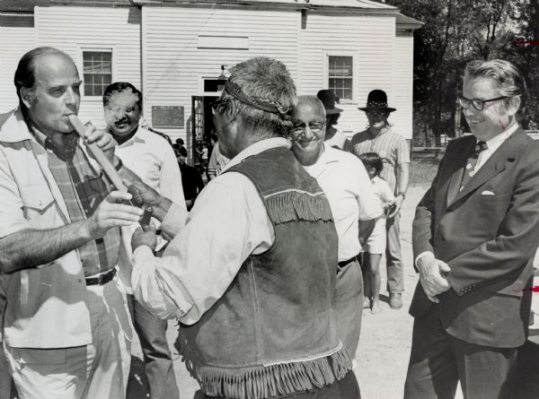 Nelson smokes a peace pipe held by Bill Baker outside a building after a meeting with tribe members. To Nelson's far right is Governor Patrick Lucey, and in the background stand several Native American men and children.