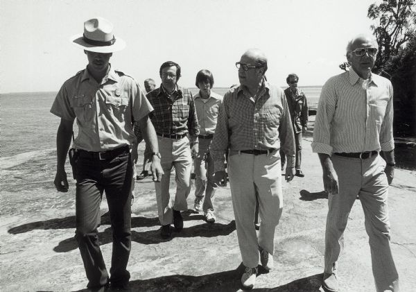 Senator Gaylord Nelson, Dave Obey, and Secretary of the Interior Cecil Andrus, walk along a beach with a park ranger during a tour of the Apostle Islands.  Immediately behind Obey is his oldest son, Craig.