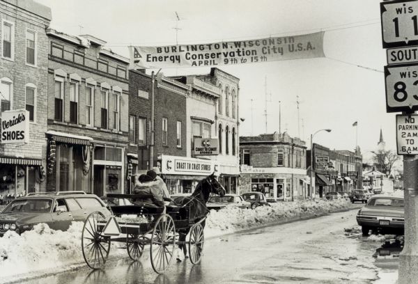The city of Burlington is visited by Gaylord Nelson during an Earth Week celebration. The photograph shows a horse-drawn carriage riding down main street, with a banner above the street advertising the city as Energy Conservation City U.S.A.