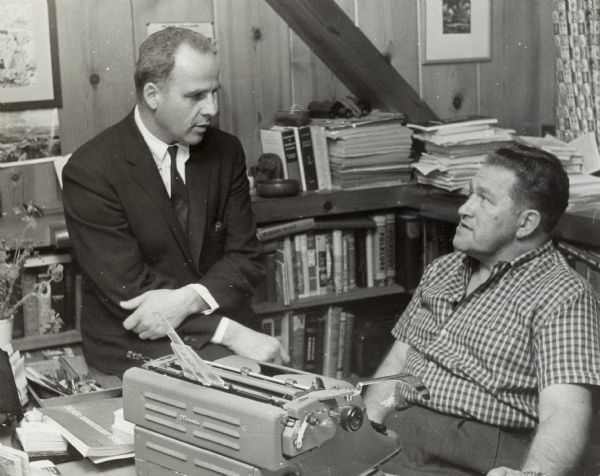 Casual portrait of August Derleth sitting behind his typewriter in his office, with Gaylord Nelson sitting on his desk to talk.
