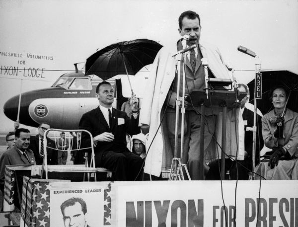 Republican presidential candidate Richard M. Nixon speaks at an outdoor event. There is an airplane in the background. Patricia Nixon is shown seated behind her husband as he speaks.