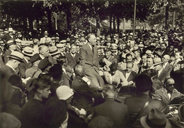 Franklin D. Roosevelt sits on the back of an automobile in the midst of a large crowd of people.