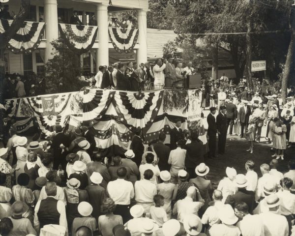 Franklin D. Roosevelt speaking from a stage to a crowd. On the stage from left to right are: Louis Howe, Governor Schmedeman (in white), Senator Robert M. La Follette, Jr. (applauding), James Hughes, Charles Broughton (?), the mayor of Green Bay (wearing a bow tie), President Roosevelt, John Roosevelt, and Senator F. Ryan Duffy.