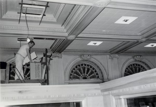 A worker on scaffolding installs ceiling tiles and new lights, concealing the plaster coffers in the library lobby of the Wisconsin Historical Society.