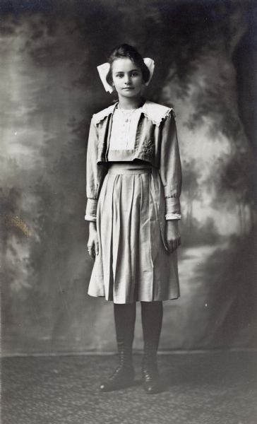 Photographic postcard of a studio portrait of Gertie Haber with a bow in her hair and her hands at her side. She wears a pleated skirt and is standing in front of a painted backdrop.