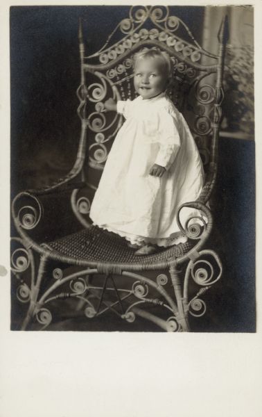 Photographic postcard of a studio portrait of an unidentified girl in a white dress standing on an ornate wicker chair. She is posed in front of a painted backdrop.