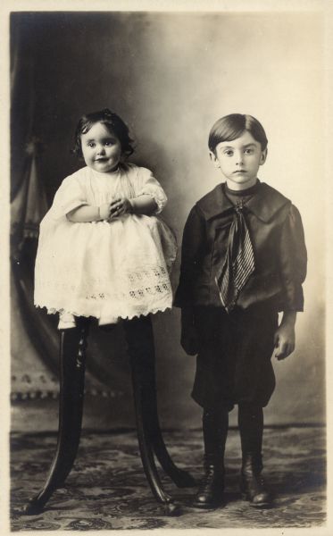 Photographic postcard of a studio portrait of two young unidentified children, perhaps a brother and sister. They are standing in front of a painted backdrop.