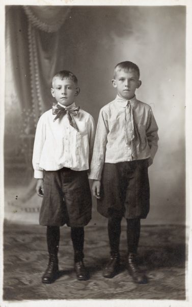 Photographic postcard of a studio portrait of two unidentified boys, perhaps brothers. They both wear neckties, light-colored shirts and dark knickerbockers (knickers), stockings and shoes. They are standing in front of a painted backdrop.