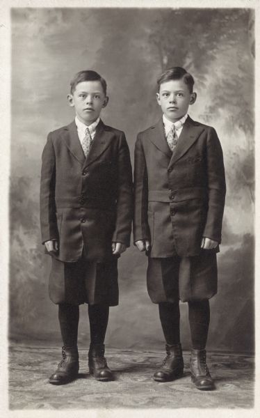 Studio portrait of, (left to right), Tony and Werner Rawls standing in front of a painted backdrop. They are wearing matching dark suits and knickerbockers (knickers).
