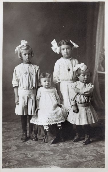 Photographic postcard of a studio portrait of four unidentified girls, perhaps family members (sisters?). They each wear a white dress, and the three older girls have large ribbons in their hair. They are standing in front of a painted backdrop.