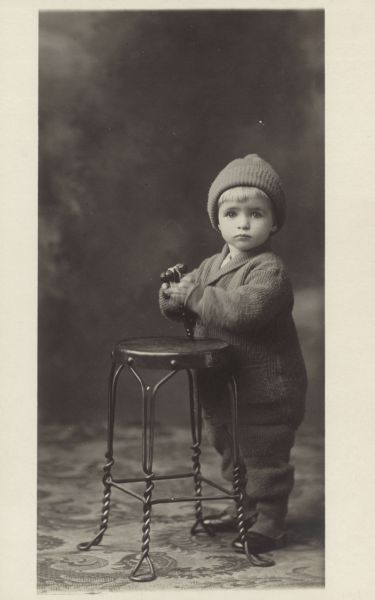 Photographic postcard of a studio portrait of an unidentified boy. He wears a hat and a woolen jumpsuit, and holds some bells in his hands. In the background is a painted backdrop.