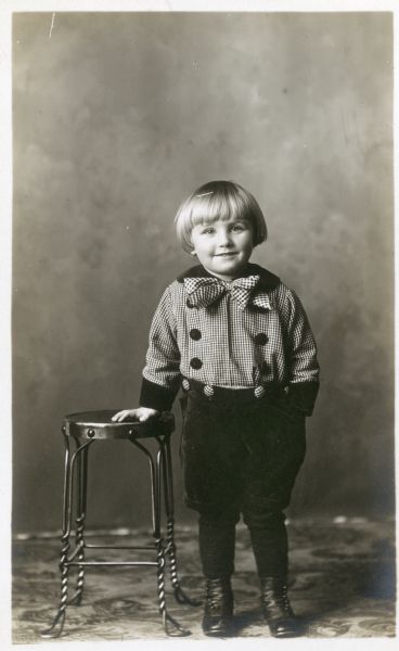 Photographic postcard of a studio portrait of an unidentified child wearing a plaid shirt and checkered bow tie, standing in front of a painted backdrop.