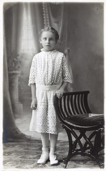 Photographic postcard of a studio portrait of a young girl in a white dress, standing in front of a painted backdrop.
