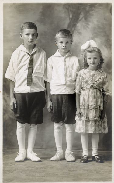 Photographic postcard of a studio portrait of three unidentified children, perhaps brothers and sister. The boys wear knickerbockers (knickers) and neckties, and the girl wears a white bow in her hair. They are standing in front of a painted backdrop.