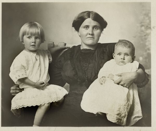 Photographic postcard of a studio portrait of an unidentified mother and her two children. The children both wear white dresses. They are posed in front of a painted backdrop.