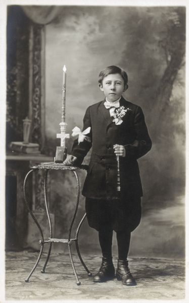Photographic postcard of a studio portrait of a boy on the occasion of his First Communion. He is dressed in jacket and knickerbockers (knickers) and holds a rosary in his left hand. He is posed in front of a painted backdrop.