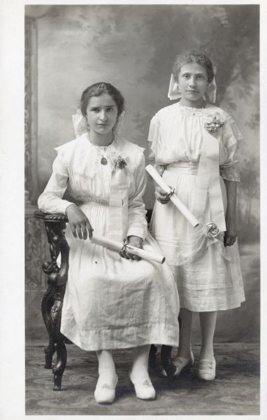 Photographic postcard of a studio portrait of two girls on the occasion of their graduation. They are dressed in white dresses with bows in their hair, corsages and they both hold rolled diplomas in their hands. They are posed in front of a painted backdrop.