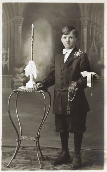 Photographic postcard of a studio portrait of an unidentified boy on the occasion of his First Communion. He is dressed in jacket and knickerbockers (knickers) and holds a rosary and catechism. There is a ribbon tied around his arm. He is posed in front of a painted backdrop.