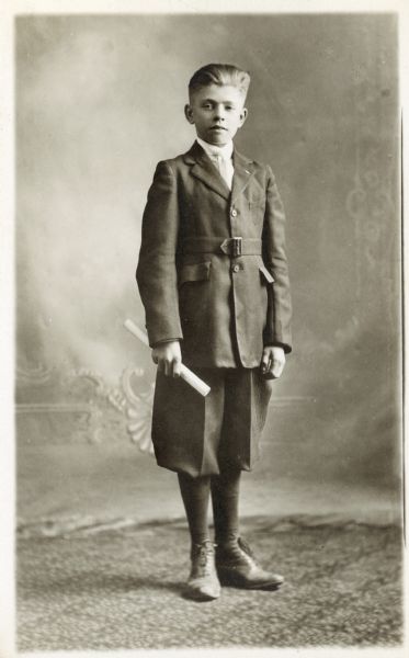 Photographic postcard of a studio portrait of a boy on the occasion of his graduation. He is dressed in jacket and knickerbockers (knickers) and holds a rolled diploma in his right hand. He is posed in front of a painted backdrop.