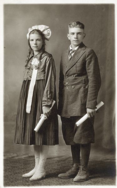 Photographic postcard of a studio portrait of Viola and Clarence Joss on the occasion of their graduation. Each of them holds a rolled diploma in their left hands. They are posed in front of a painted backdrop.