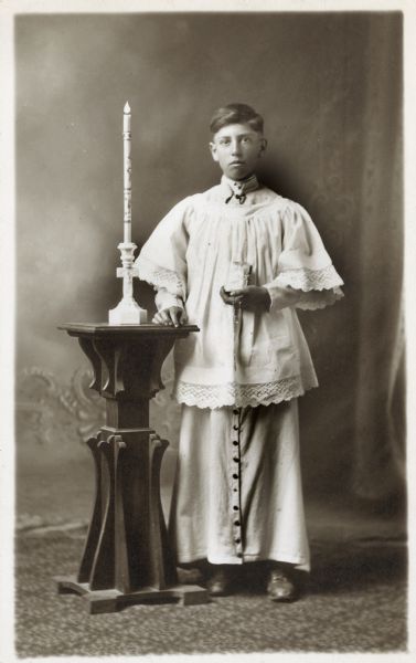Photographic postcard of a studio portrait of Joe Dahman on the occasion of his First Communion. He is dressed in church robes and holds a catechism and rosary in his left hand, and is standing next to a lighted candle. He is posed in front of a painted backdrop.