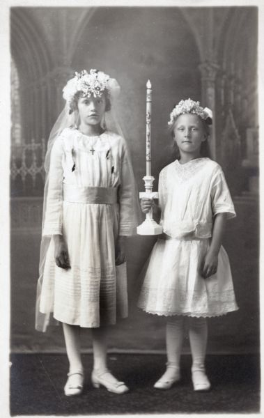 Photographic postcard ofa studio portrait of two girls on the occasion of their First Communion. They both wear white dresses and decorative headpieces with flowers. The girl on the left is wearing a long veil, and the girl on the right holds a cross-shaped candle-holder in her right hand. They are posed in front of a painted backdrop.
