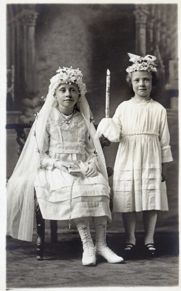 Photographic postcard of a studio portrait of two girls on the occasion of their First Communion. They both wear white dresses and decorative headpieces with flowers. The girl on the left, sitting in a chair, is wearing a veil and holds a catechism and rosary in her right hand. The girl on the right is holding a candle in her right hand. They are posed in front of a painted backdrop.