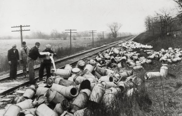 Milk strikers and a pile of empty milk cans next to railroad tracks. Bill Nemeth (left) is pouring out milk in an attempt to create a shortage that would raise milk prices.