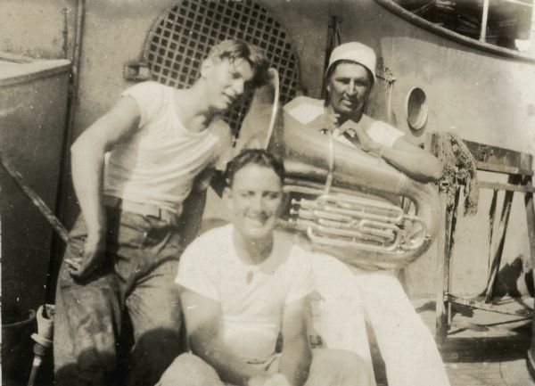 Candid portrait of three men in white t-shirts. Angus Lookaround, in a white hat, is seated, holding a tuba. The men appear to be aboard a ship, possibly the USS <i>New Hampshire</i>, during Lookaround’s service in the United States Navy.” 
